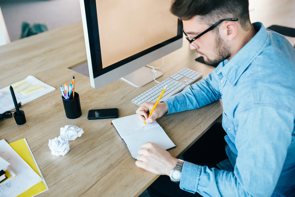 Young attractive man in glassess is working at his workplace in office. He wears blue shirt. He is writing in notebook. View from above.